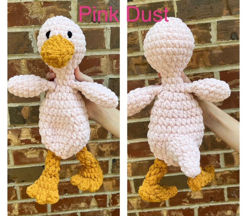 May include: Pink crocheted duck plush toy with yellow feet and beak. The duck is standing on a brick wall.
