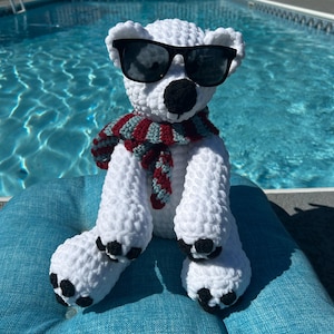May include: A handmade white crochet polar bear wearing black sunglasses and a red, gray, and green scarf. The bear has black paws and a black nose. It is sitting on a blue cushion near a swimming pool.