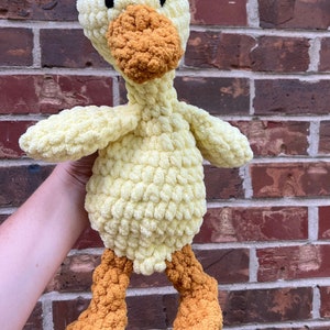 May include: A crocheted yellow duck plush toy with orange feet and beak. The duck is standing on a brick wall.