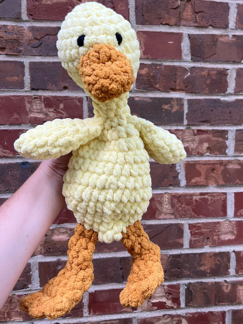 May include: A crocheted yellow duck plush toy with orange feet and beak. The duck is standing on a brick wall.
