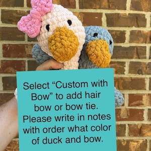 May include: Two crocheted duck plushies, one with a pink bow. The sign reads "Select "Custom with Bow" to add hair bow or bow tie. Please write in notes with order what color of duck and bow."