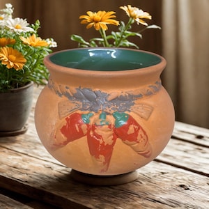 May include: A terracotta-colored ceramic planter with a green interior. The planter features a design of three red chili peppers with green stems. The planter is sitting on a wooden surface with flowers in the background.