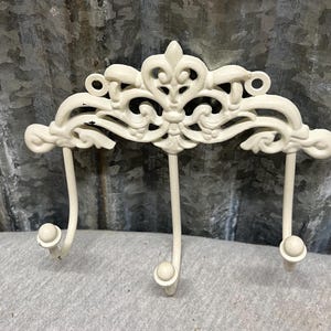 May include: Off-white, ornate metal wall hook with three curved hooks. The decorative top features a fleur-de-lis design and scrollwork. The hooks are topped with small, round knobs. The background is a corrugated metal sheet.