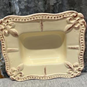 May include: A rectangular cream-colored ceramic tray with a raised border featuring a beaded edge and fleur-de-lis motifs at each corner. The piece has a distressed finish with brown accents highlighting the decorative relief details.