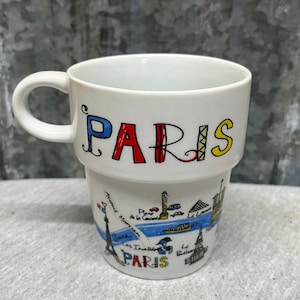 May include: White ceramic mug with the word "PARIS" in colorful letters. The mug features a handle and a lower section with illustrations of Parisian landmarks, including the Eiffel Tower and the Seine River.