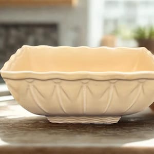May include: A cream-colored, rectangular ceramic serving bowl with a scalloped rim and decorative embossed details. The bowl has a slightly raised base with a wavy pattern. The background is blurred, suggesting a kitchen setting.