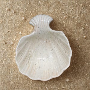 May include: A white ceramic seashell-shaped dish with a speckled pattern. The dish has a scalloped edge and a handle-like extension. The background is a textured tan fabric with small shell fragments.