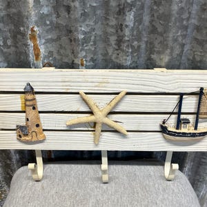 May include: A white wooden wall-mounted rack featuring three metal hooks. The board is decorated with a tan lighthouse figurine on the left, a central beige starfish, and a small blue and white sailboat model on the right side of the panel.