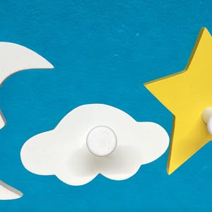 May include: Wooden wall hooks in the shape of a crescent moon, cloud, and star. The moon and cloud are white, the star is yellow. Each has a knob for hanging items. The background is a blue color.