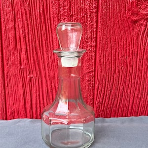 May include: A clear glass bottle with a stopper. The bottle has a rounded base and a square-shaped body. The bottle is sitting on a gray surface.