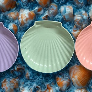 May include: Three seashell-shaped dishes in pastel colors: lavender, mint green, and coral. The dishes have a ribbed texture and are arranged on a background of seashells.