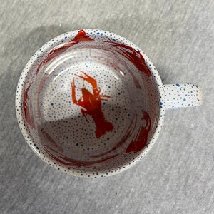 May include: White ceramic mug with a red crawfish design inside. The mug has a handle and is decorated with blue polka dots on the rim and handle. Red paint splatters are visible inside the mug.
