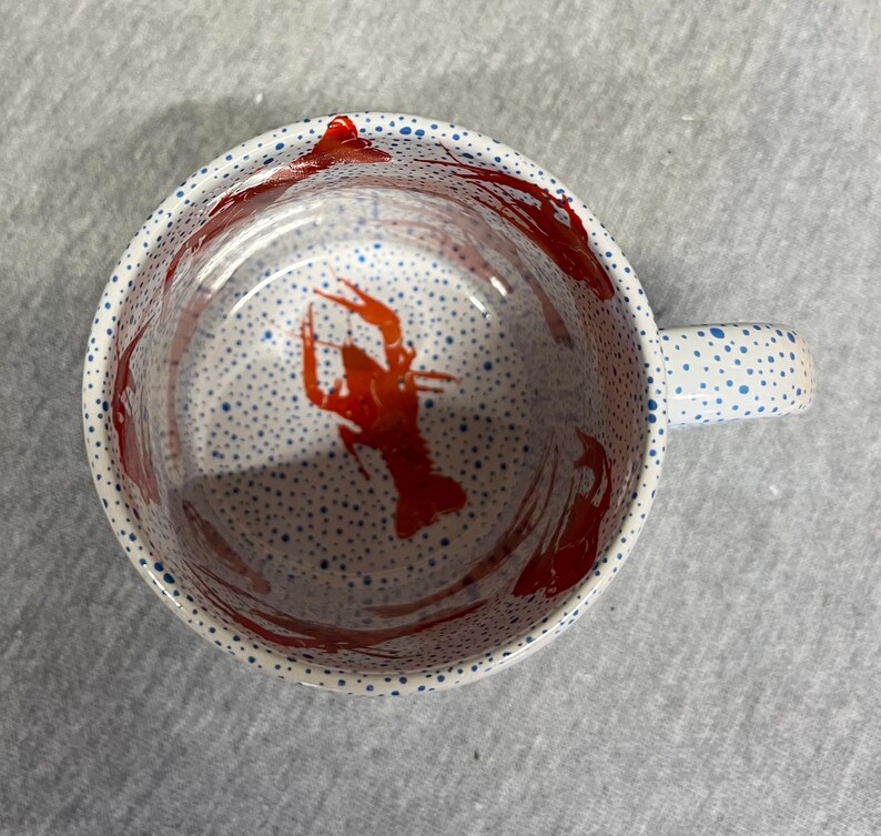 May include: White ceramic mug with a red crawfish design inside. The mug has a handle and is decorated with blue polka dots on the rim and handle. Red paint splatters are visible inside the mug.