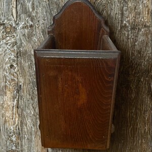 May include: A wall-mounted wooden storage bin featuring a dark brown stained finish with a visible wood grain pattern. The container has a rectangular body with a tall, arched back panel and an open top, designed for holding various small items.