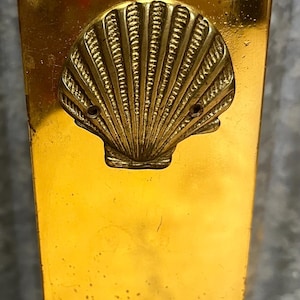 May include: A vintage brass wall-mounted match holder. The rectangular holder features a detailed seashell design. The brass has a golden hue with some signs of wear, adding to its antique charm.