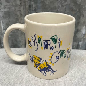 May include: Cream-colored ceramic mug with the words "MARDI GRAS" in blue, green, and yellow. The mug features a yellow and blue mask design. The mug has a curved handle.