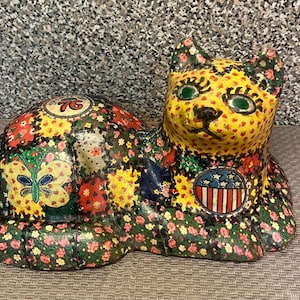 May include: A decorative cat figurine featuring a patchwork-style design with various floral patterns in red, yellow, and green. The cat displays a yellow face with green eyes, a butterfly graphic, a circular 76 emblem, and a red, white, and blue shield.