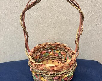 May include: A small, round woven basket with a pink handle. The basket is made of natural materials and has a colorful, geometric pattern.