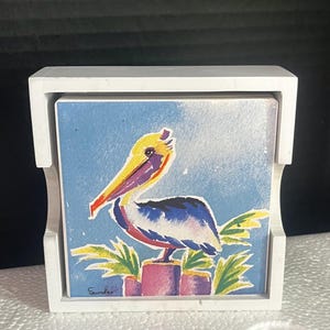 May include: A square coaster featuring a watercolor-style illustration of a pelican perched on a post against a blue background, housed in a white wooden holder. The artwork includes green foliage accents and a signature in the bottom left corner.
