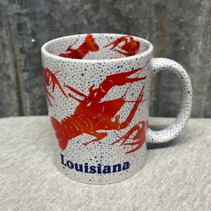 May include: White ceramic mug with a red crawfish design and the word "Louisiana" in blue. The mug has a speckled pattern of blue dots and a curved handle.