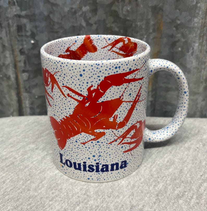 May include: White ceramic mug with a red crawfish design and the word "Louisiana" in blue. The mug has a speckled pattern of blue dots and a curved handle.