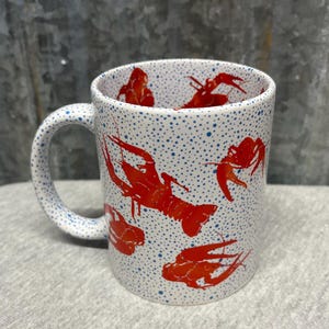 May include: White ceramic mug with a handle, featuring a pattern of red crawfish and blue polka dots. The crawfish are in various poses, adding visual interest to the design. A fun and unique kitchenware item.