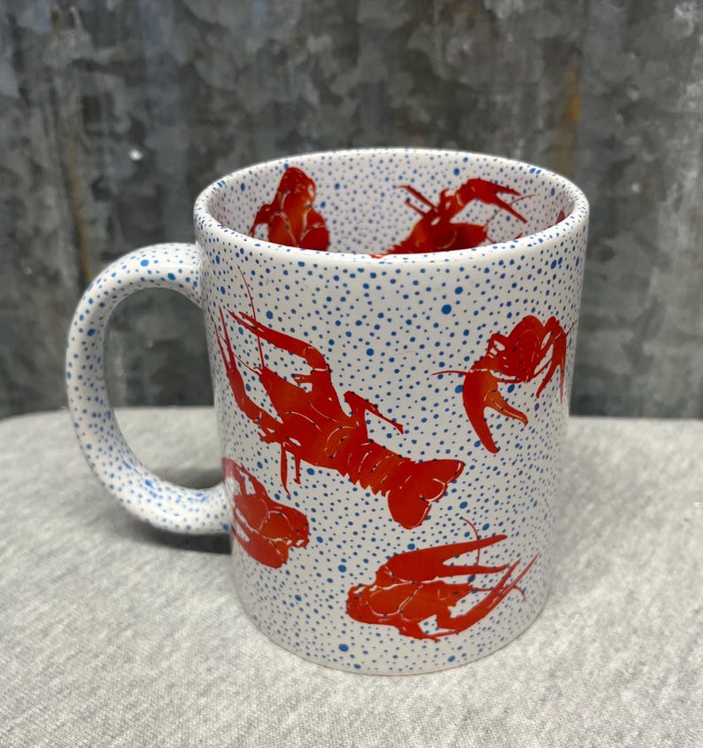 May include: White ceramic mug with a handle, featuring a pattern of red crawfish and blue polka dots. The crawfish are in various poses, adding visual interest to the design. A fun and unique kitchenware item.