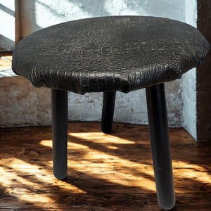 May include: A small, black stool with a round seat and three legs. The seat has a textured, crocodile-like pattern. The legs are cylindrical and painted black. The stool is set on a wooden floor.