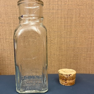 May include: A clear glass bottle with a square base and a rounded top, accompanied by a cork stopper. The bottle has embossed text that reads "POUND PURE ONE". The bottle and stopper are on a blue surface.