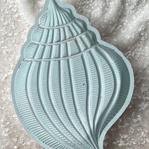 May include: A light blue seashell-shaped decorative dish. The dish has a textured surface with detailed ridges and a slightly distressed finish. The seashell is a light blue color and sits on a textured, off-white surface. The dish is approximately 6 inches long.