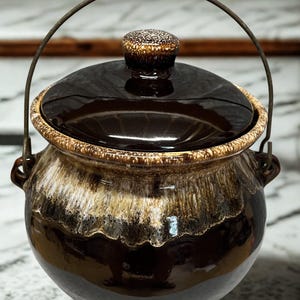 May include: A vintage brown ceramic pot with a lid and a metal handle. The pot has a glossy finish with a speckled lid and a two-tone design of brown and beige. The pot has a round shape and small feet.