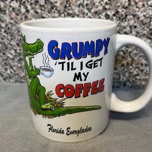May include: A white ceramic mug featuring a green alligator illustration holding a steaming cup of coffee. The text reads GRUMPY 'TIL I GET MY COFFEE in blue and red letters, with Florida Everglades printed in black script at the bottom of the mug.