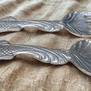 May include: A silver-colored fork and spoon set. The handles are shaped like fish, with detailed scales and fins. The spoon has a rounded bowl, and the fork has three tines. The set is designed for serving.