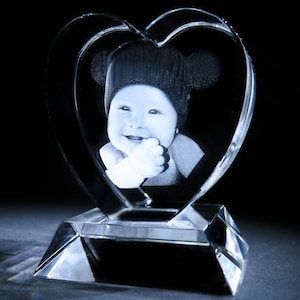Puede incluir: Un pisapapeles de cristal en forma de coraz&oacute;n con una imagen grabada en 3D de un beb&eacute; con un sombrero. El pisapapeles tiene una base de cristal transparente.