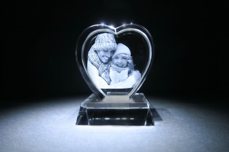 Puede incluir: Un pisapapeles de cristal en forma de coraz&oacute;n con una imagen grabada en 3D de una pareja con ropa de invierno.