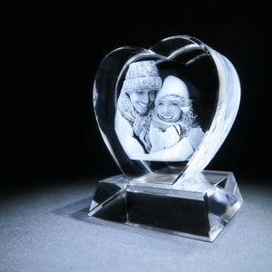 Puede incluir: Un pisapapeles de cristal en forma de coraz&oacute;n con una imagen grabada en 3D de una pareja con ropa de invierno. El pisapapeles est&aacute; sobre una base de cristal transparente.