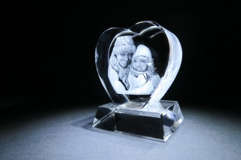 Puede incluir: Un pisapapeles de cristal en forma de coraz&oacute;n con una imagen grabada en 3D de una pareja con ropa de invierno. El pisapapeles est&aacute; sobre una base de cristal transparente.