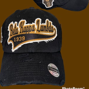 May include: Black distressed baseball cap with embroidered "Beta Kappa Lambda 1939" text in gold and white. Features a distressed brim and a small patch with a pharaoh design. The cap has a vintage look.