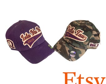 Könnte beinhalten: Zwei Baseballkappen im Used-Look. Eine ist lila mit dem Text Iota Mu Nu 2015 und einem Omega-Symbol. Die andere hat ein Tarnmuster mit dem Text Delta Tau 1958 und VasOmega. Beide Kappen haben ausgefranste Kanten und aufgestickte Applikationen.