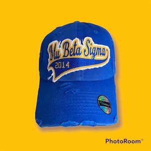 Könnte beinhalten: Königsblaue Baseballkappe mit Used-Details. Die Kappe hat einen gestickten Aufnäher mit dem Text "Mu Beta Sigma 2014" in Weiß und Gold. Auf der Seite befindet sich ein Vintage-Aufkleber.
