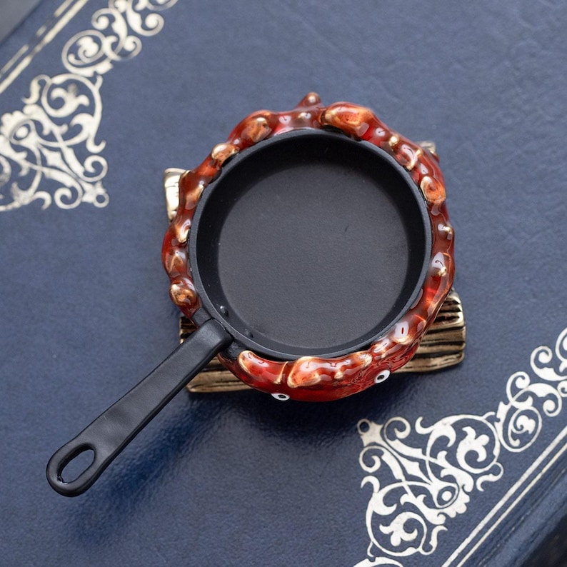 May include: A miniature black frying pan with a long handle, set within a decorative crab-shaped frame. The crab is red and brown with small white eyes. The pan rests on a small, gold-colored base, all set against a dark blue background.