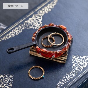 May include: A decorative ring dish shaped like a miniature frying pan. The dish is black with a red and brown crab-like border. Two gold rings are inside the pan, and a gold ring with a turquoise stone sits on the surface.