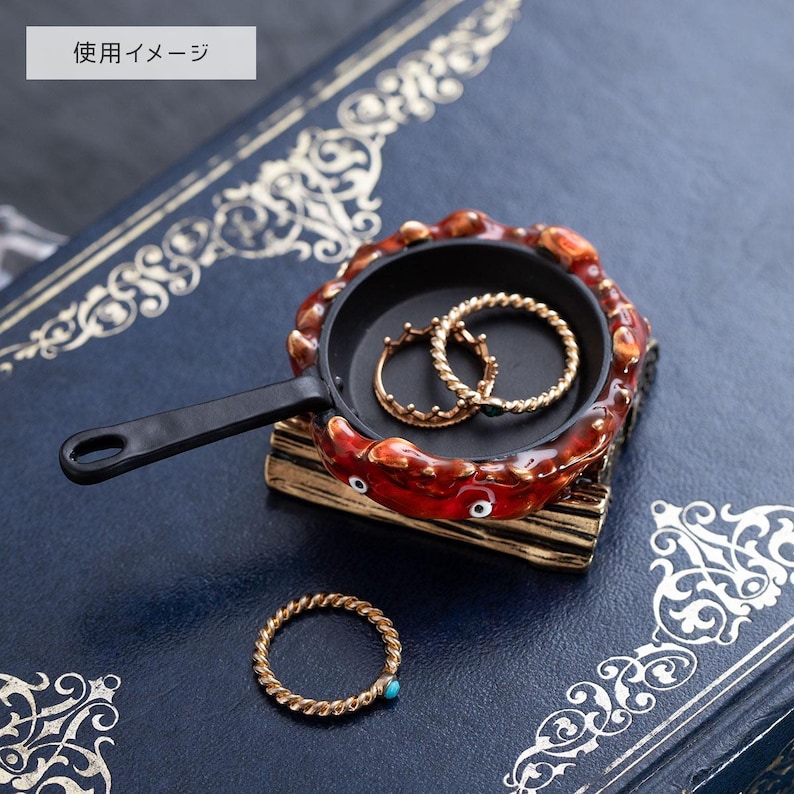 May include: A decorative ring dish shaped like a miniature frying pan. The dish is black with a red and brown crab-like border. Two gold rings are inside the pan, and a gold ring with a turquoise stone sits on the surface.