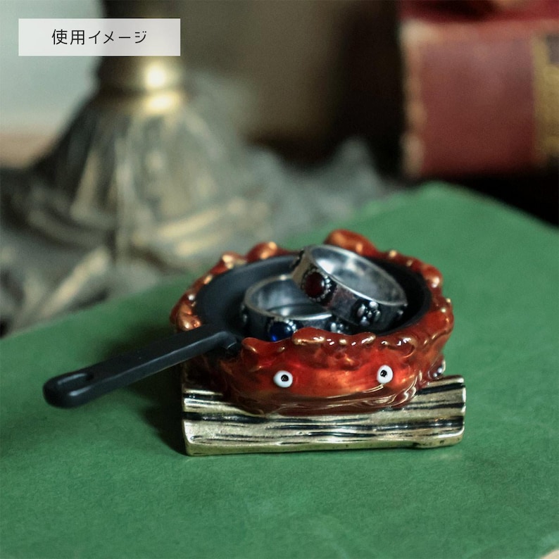 May include: A whimsical ring holder shaped like a red crab with a black pan. Two silver rings with gemstones rest inside. The crab sits on a wooden base. The handle is black.