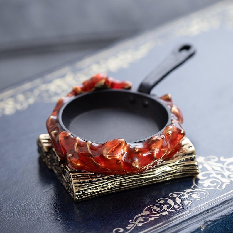 May include: A miniature black frying pan with a black handle, set within a decorative, fiery red and gold resin frame. The pan rests on a gold-trimmed, wood-like base, creating a unique kitchen decor item.