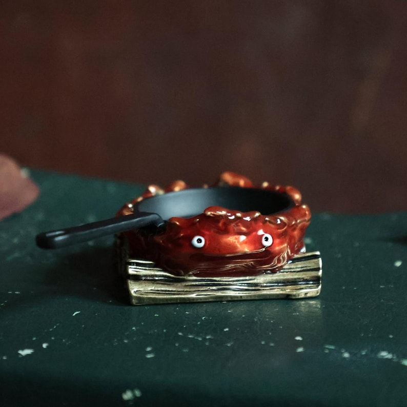 May include: A miniature, whimsical, red and black pan with a handle, resting on a wooden base. The pan has two white eyes, giving it a cartoonish appearance. The base is gold-colored.
