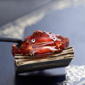 May include: A red and gold crab-shaped incense burner with white eyes. The crab sits on a rectangular base with gold trim. The burner is held by a black handle, and steam is visible.