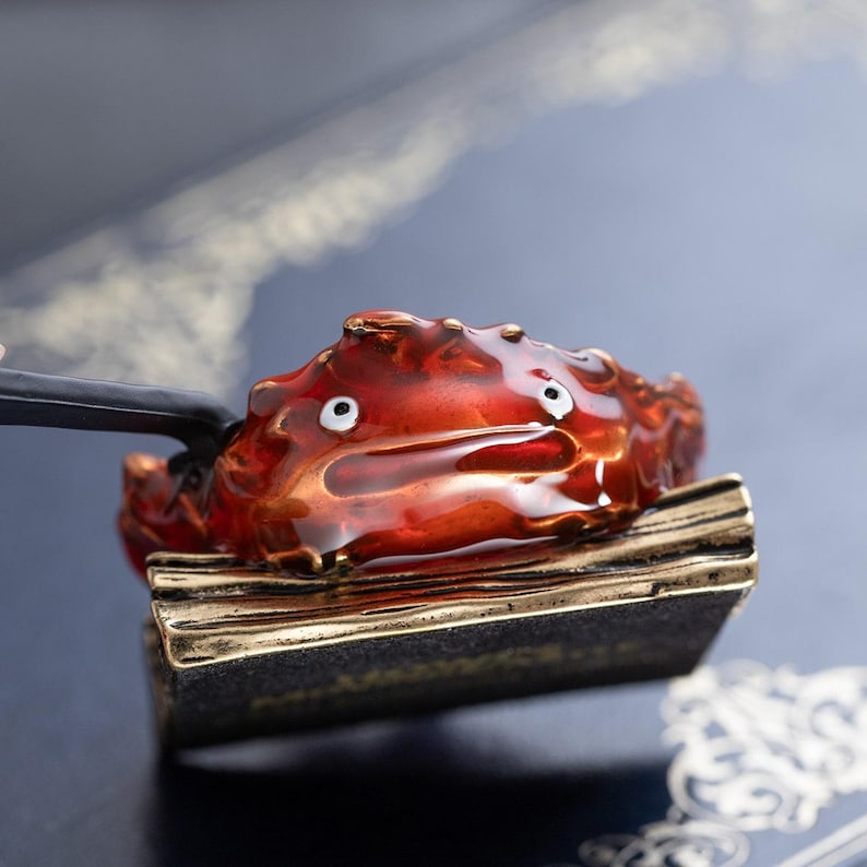 May include: A red and gold crab-shaped incense burner with white eyes. The crab sits on a rectangular base with gold trim. The burner is held by a black handle, and steam is visible.