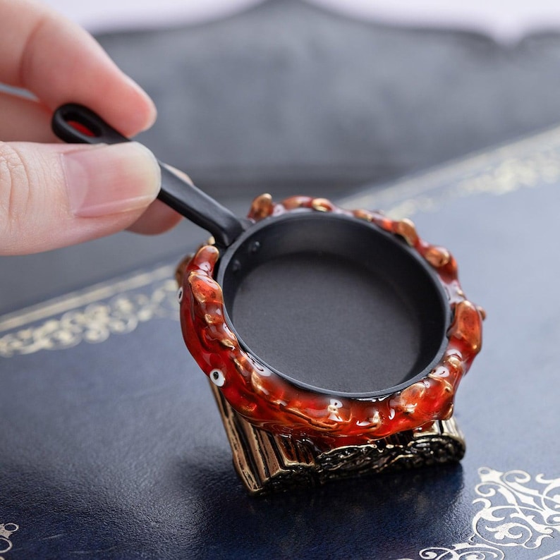 May include: Miniature black frying pan with a red and gold decorative base. The base has a crab-like design with a detailed, textured appearance. The pan is held by a hand against a blue background.