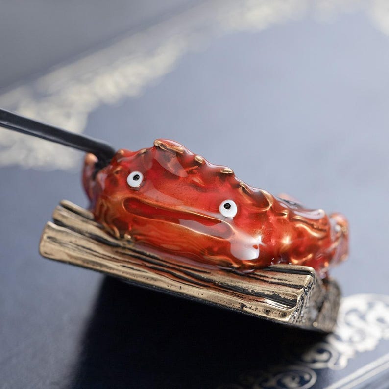 May include: A red and gold crab-like figurine with white eyes, perched on a wooden base. The figurine has a glossy finish and is attached to a black rod. The base is rectangular with a gold-colored edge. The overall design is whimsical.